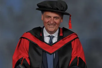 Steven Moffat smiling at the camera while wearing a PhD cap and gown in front of a blue backdrop