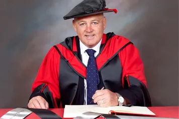 Warren Gatland smiles at the camera with a book on a red desk while wearing a PhD cap and gown in front of a blue backdrop
