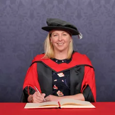 Honorary fellow Jayne Ludlow smiles at the camera while writing in a large book on a red desk and wearing a PhD cap and robe