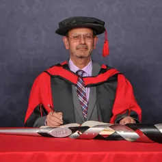 Honorary fellow Keshav Singhal looks at the camera as they write in a large book on a red desk while wearing a PhD cap and gown