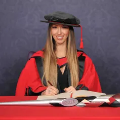 Honorary fellow Sabrina Cohen smiles at the camera as they write in a large book on a red desk while wearing a PhD cap and gown