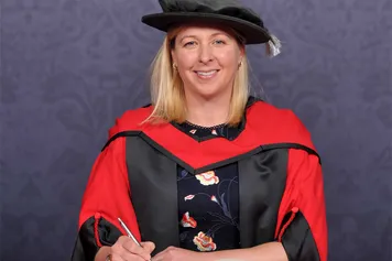 Jayne Ludlow smiles at the camera while wearing a doctorate's cap and gown in front of a blue backdrop