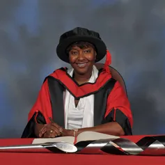 La Chun Lindsay writes in a large book next to a ceremonial mace on a red desk while wearing a doctorate's cap and gown in front of a blue backdrop.