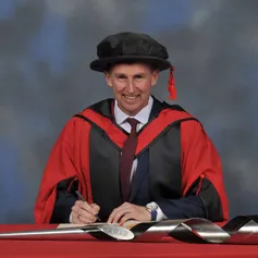 Nigel Owens writes in a large book next to a ceremonial mace on a red desk while wearing a doctorate's cap and gown in front of a blue backdrop.