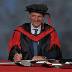 Steven Moffat writes in a large book next to a ceremonial mace on a red desk while wearing a doctorate's cap and gown in front of a blue backdrop.