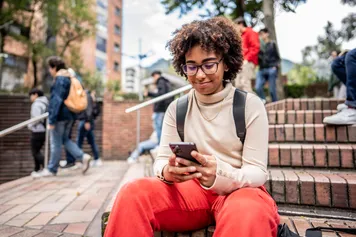 A student sitting outside campus using mobile phone and smiling