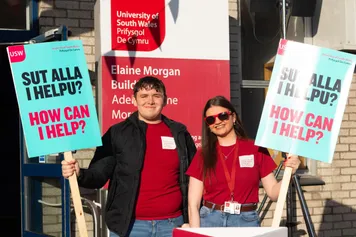 Two student ambassadors smiling at the camera during an open day, holding signs that read 'how can I help?'