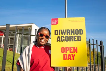 A student ambassador smiling in the sun, wearing red USW sunglasses and holding an Open Day sign