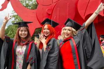 Three students dressed in graduation robes and graduation caps smile and pose in front of the red dragon outside Newport's ICC