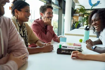 A group of students laugh and chat while sat around a table in a study zone at Newport campus