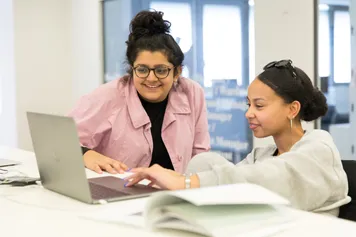 Two students sat at a table working on a laptop.