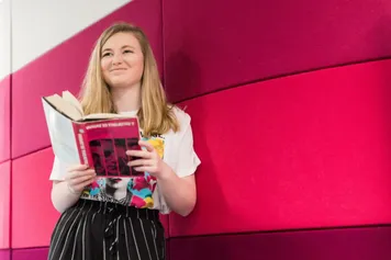 A student standing against a pink background in the Cardiff campus library holding a book and smiling