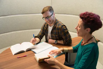 Two people studying together at a table, surrounded by books and papers.