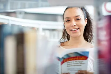 A student smiling and looking through a bookshelf in the library.