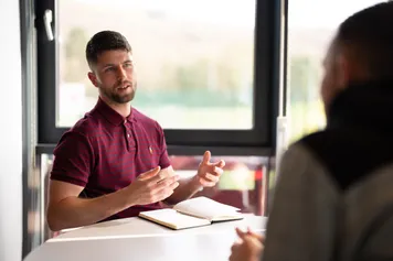 A student talking whilst sat at a table with a book in front of him.