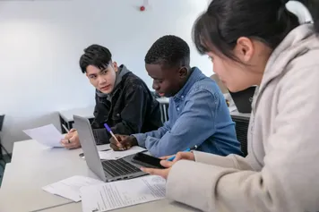 Three students sitting at a table engaged in conversation, with laptops and papers in front of them.