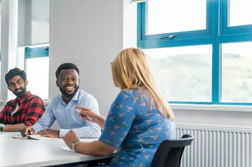 Three students sat at a table studying together with windows in the background. One of the students is pointing and the other two are listening and smiling.