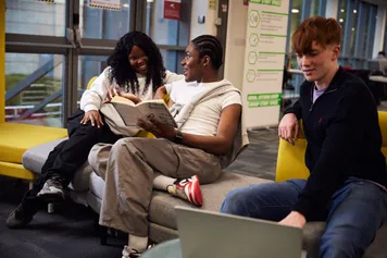 Three students studying together. Two are looking at a book and the other is using a laptop.