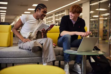 Two students looking at a book and using a laptop in the library.