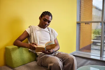 A student sat on a small sofa chair in front of a window in the library reading a book.