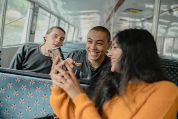 Three students are sat together on a bus. One of them is turning around to show the other two her mobile phone and they are laughing.