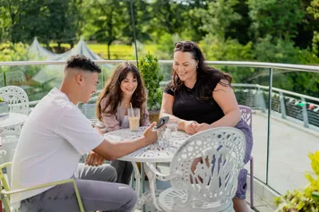 Three students are sat together outside a cafe with drinks. There is a park with lots of greenery behind them.