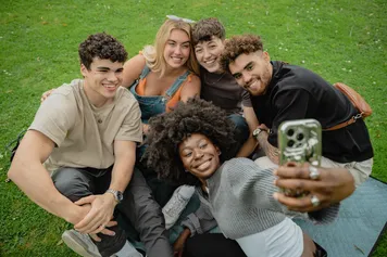 A group of students are sat together on the grass, taking a selfie.