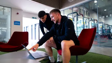 Two men having a conversation whilst looking at a laptop in the sports park