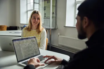 two accounting and finance students sat at a table, both at laptops, engaging in conversation