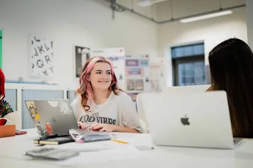 A student with pink and brown hair is sitting in a studio, working on a laptop and smiling.