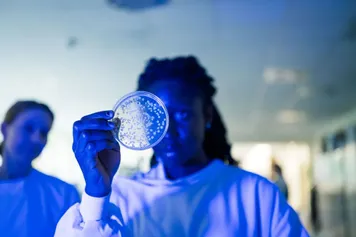 Two students in lab coats proudly hold up a petri dish together in a science lab setting.