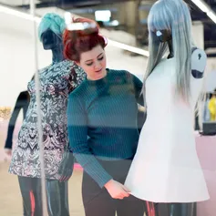A student adjusts a white dress on a black mannequin which is wearing a light silvery blue wig in a shop window