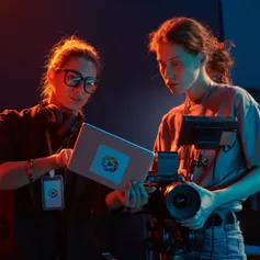 A director points at a tablet screen while talking to a person with a camera on a darkly lit film set