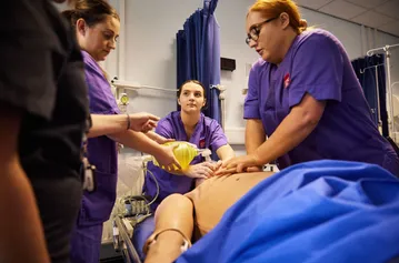 Students in scrubs are performing a CPR routine on a dummy in a hospital ward setting.