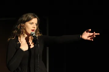 A person dressed in black, is on a black stage singing into a microphone while reaching out to the audience.