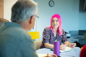 A student with shoulder-length bright pink hair listens intently to a fellow student while taking notes in a practice counselling session