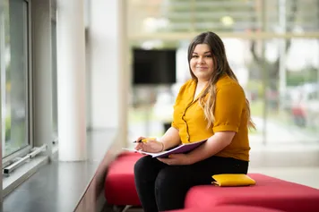 A student is sat looking into the camera, they are holding an open notebook and a pen in the other hand.