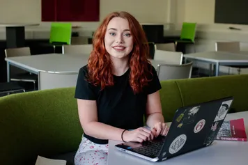 Sociology student Sara Daniels smiles at the camera while working on a laptop from a green sofa
