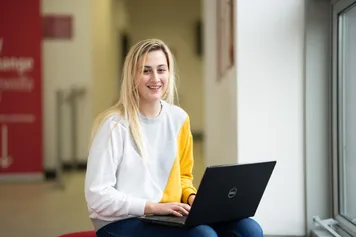 A student is smiling into the camera while holding a laptop on their knee.