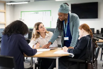 3 college students sat around a table working with a teacher leaning over to help
