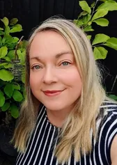 Bethan Rees  yn sefyll yn yr awyr agored o flaen planhigion gwyrddion, yn gwisgo top streipiog du a gwyn