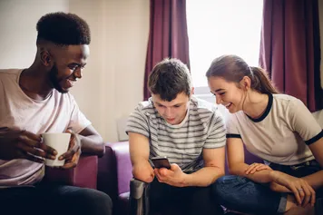Three diverse friends laugh while looking at a smart phone sat on sofas in student accommodation