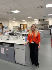 Hannah Seale yn sefyll mewn labordy gwyddoniaeth, yn pwyso ar fainc waith gyda chyfarpar a gwydrwaith ar hyd yr ystafell.