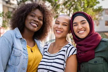 A close-up of three women, who all belong to the global majority, posing while smiling at the camera together