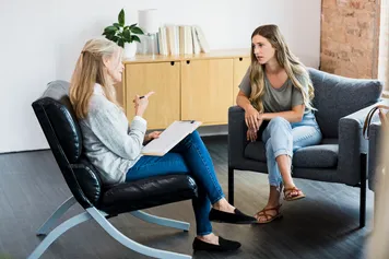 A young person sat in a chair talks to an older adult in a chair opposite in a therapy setting