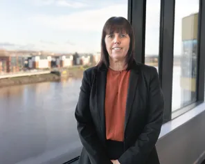 Claire Parmenter yn sefyll yn gwenu ar y camera o flaen ffenestr gydag afon y tu ôl iddi