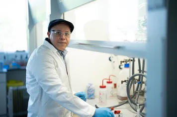 A chemistry researcher looks at the camera while wearing a white laboratory coat and blue gloves as they carry out an experiment in a chemistry laboratory