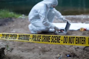 A crime scene investigator dressed in full white overalls looks for tools in a silver case at an active crime scene near a river cordoned off by yellow crime scene tape
