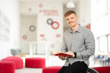 PhD student Iwan Rowlands smiles at the camera while holding an open red hardback book and stood in the foya of the South Wales Business School at the Treforest campus with red sofas in the background