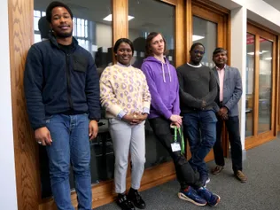 A group of five students of various ages smile while standing in front of a meeting room in the Postgraduate Centre at the USW Treforest campus